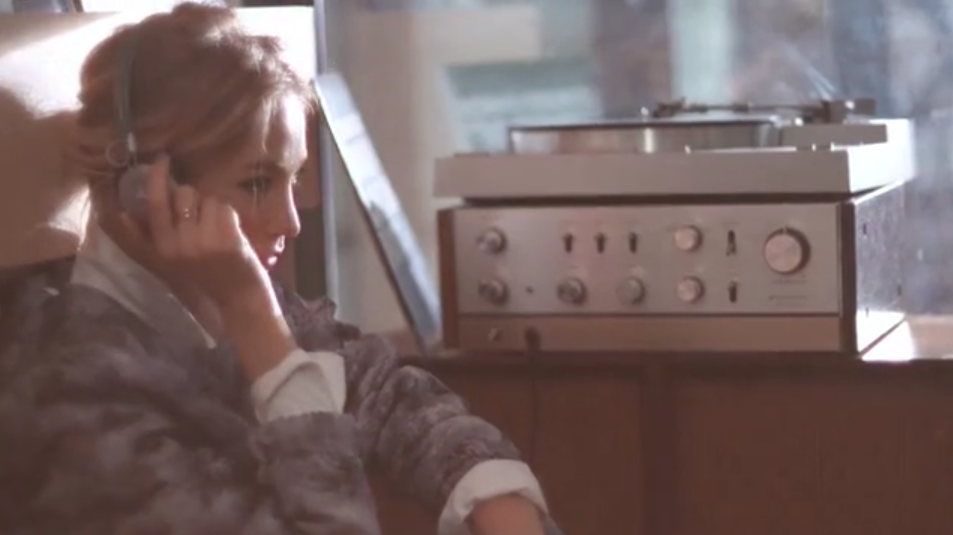 Blonde woman with headphones sits indoors listening to a silver-colored record player on a wooden shelf, warm lighting.