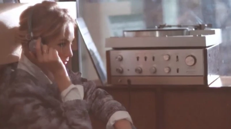Blonde woman with headphones sits indoors listening to a silver-colored record player on a wooden shelf, warm lighting.