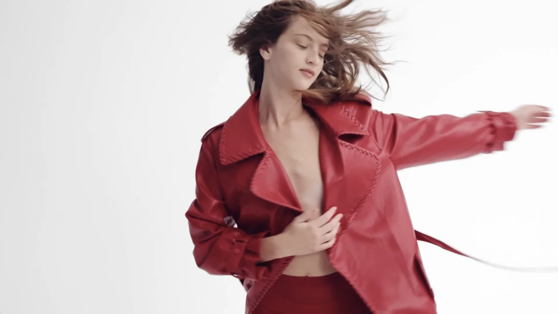Woman in motion wearing a red leather trench coat and matching bottoms against a white background, capturing a blend of fashion and simplicity.