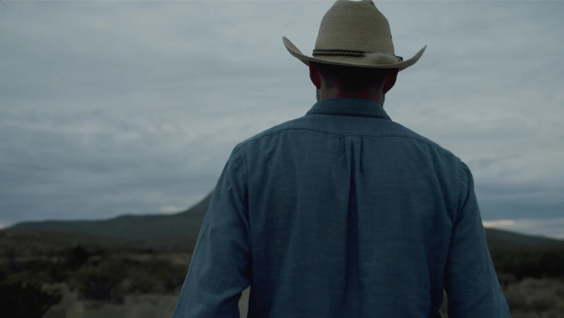 Back view of adult male wearing cowboy hat and blue shirt in rural setting with cloudy sky and mountain in the distance.