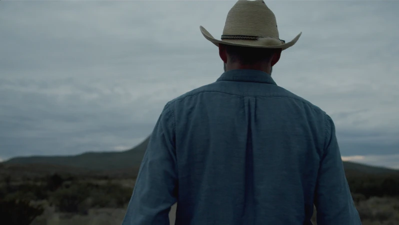 Back view of adult male wearing cowboy hat and blue shirt in rural setting with cloudy sky and mountain in the distance.
