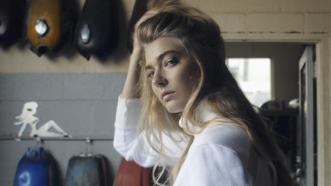 Indoor portrait of a fair-skinned, blonde woman in a white turtleneck sweater, posing with a neutral expression, with a neutral background.