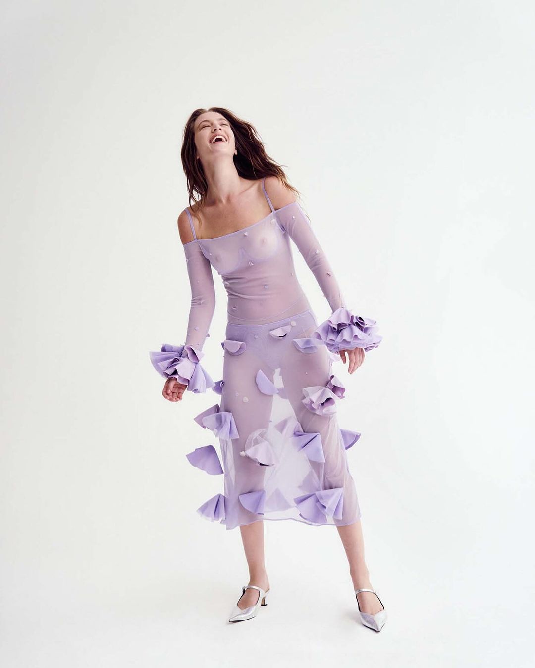 A full shot of a smiling adult woman in a studio. She is wearing a lilac sheer dress with floral accents and matching heels.