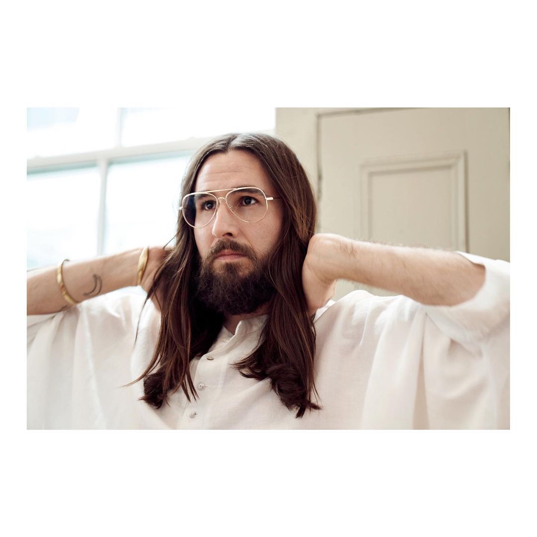 Portrait of a bearded adult male wearing wire-frame glasses and a white shirt indoors, hands behind his head, medium shot, natural light.