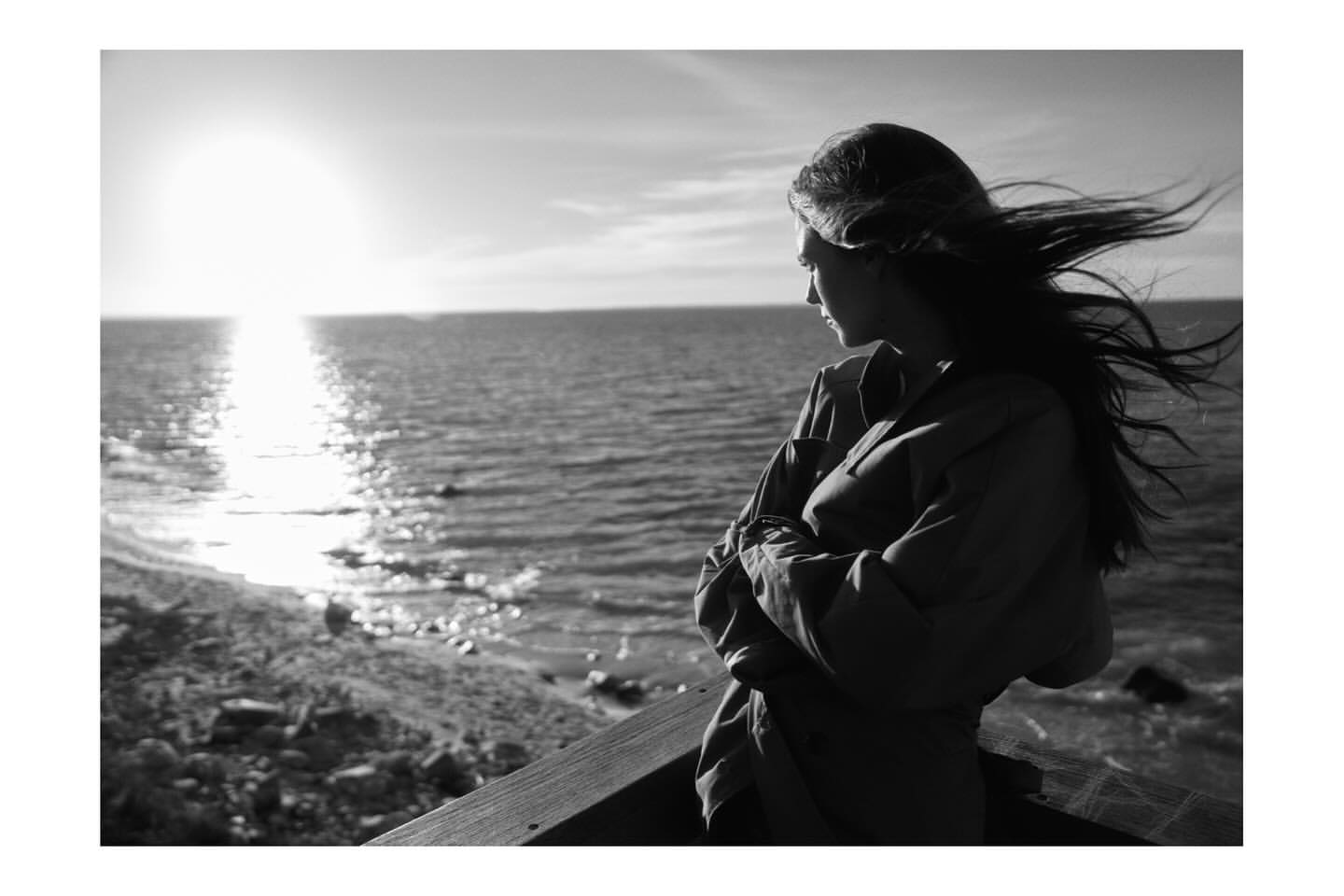 Black and white portrait of a woman with long hair blowing in the wind looking out at the ocean at sunset.