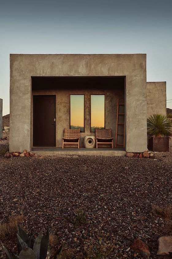 Exterior of a modern minimalist building with a recessed porch featuring chairs, a door, and reflections of the outside landscape.