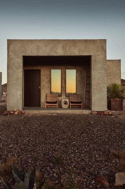 Exterior of a modern minimalist building with a recessed porch featuring chairs, a door, and reflections of the outside landscape.