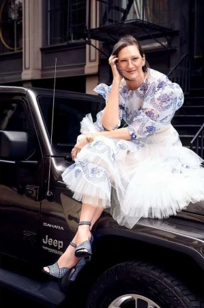 Woman in white dress with blue floral pattern sits on Jeep Wrangler in urban setting with brown buildings in the background, stylish pose.