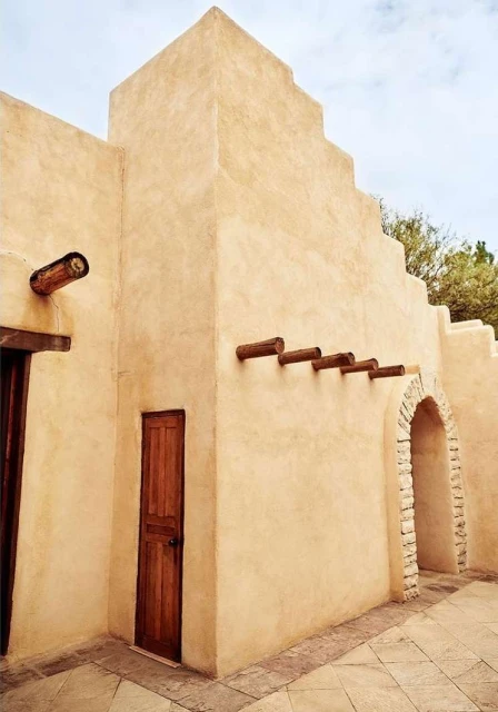 Exterior shot of a tan stucco building with wooden accents, featuring an arched entrance and a small wooden door, under a bright sky.