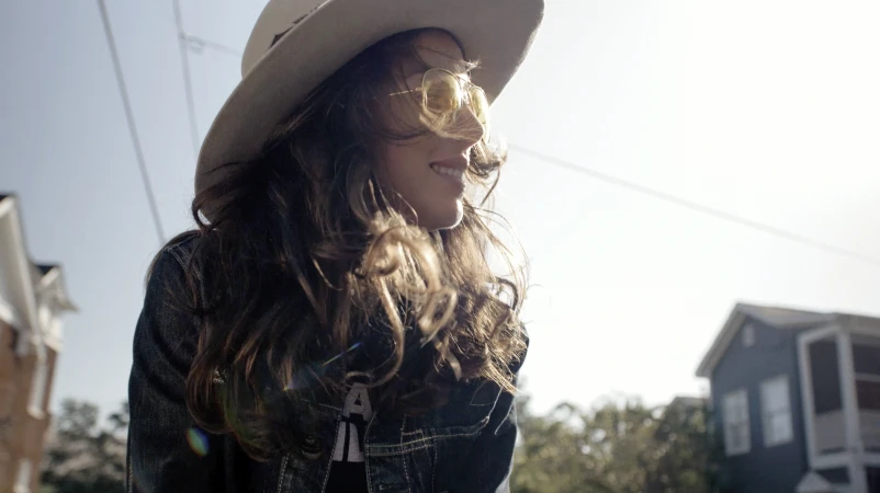 Woman wearing yellow tinted sunglasses and a wide brimmed hat outdoors by houses