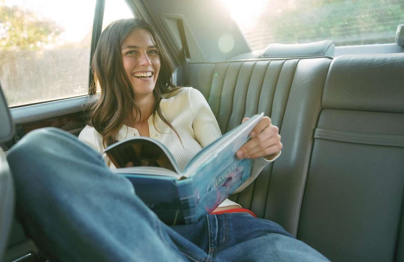 Woman smiles and reads magazine while sitting in the backseat of a car with a leather interior, illuminated by natural light.