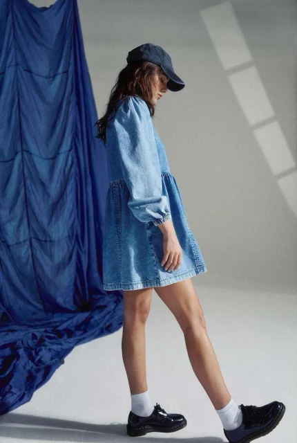 A young woman in a denim dress and baseball cap walks against a blue-draped backdrop, her modern, casual style highlighted in the studio shot.