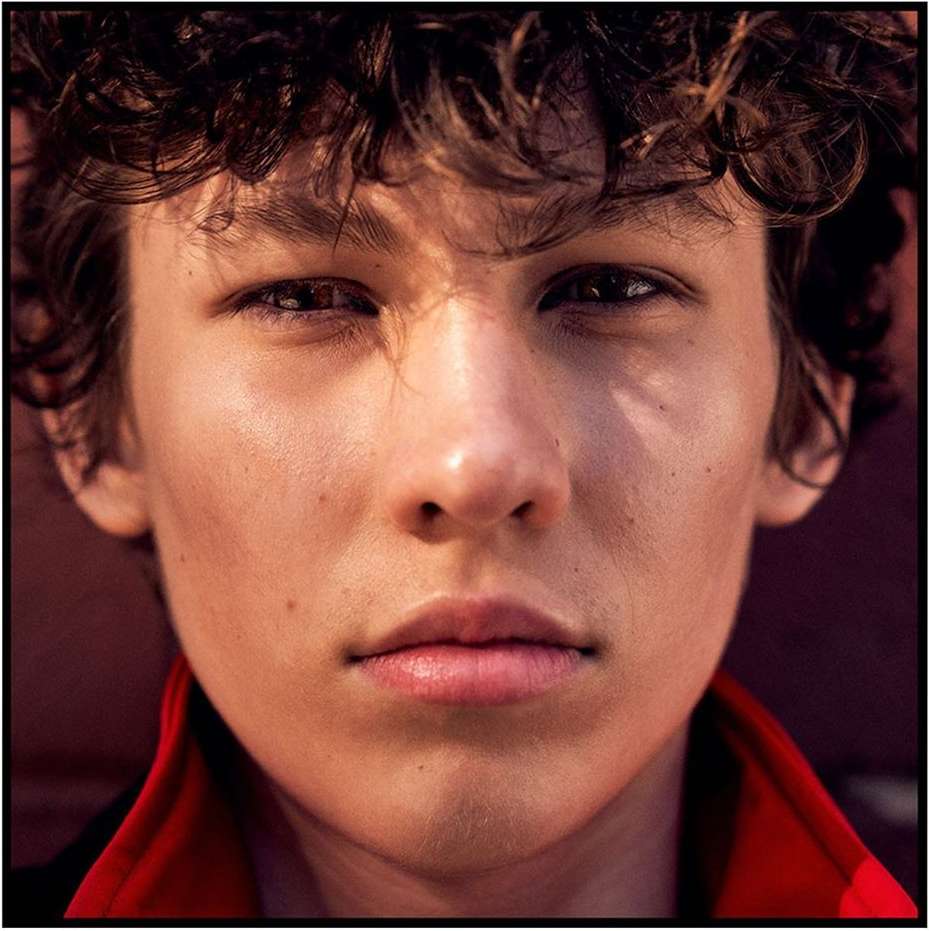 Close-up portrait of a young man with curly dark hair wearing a red collar against a dark background