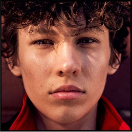 Close-up portrait of a young man with curly dark hair wearing a red collar against a dark background