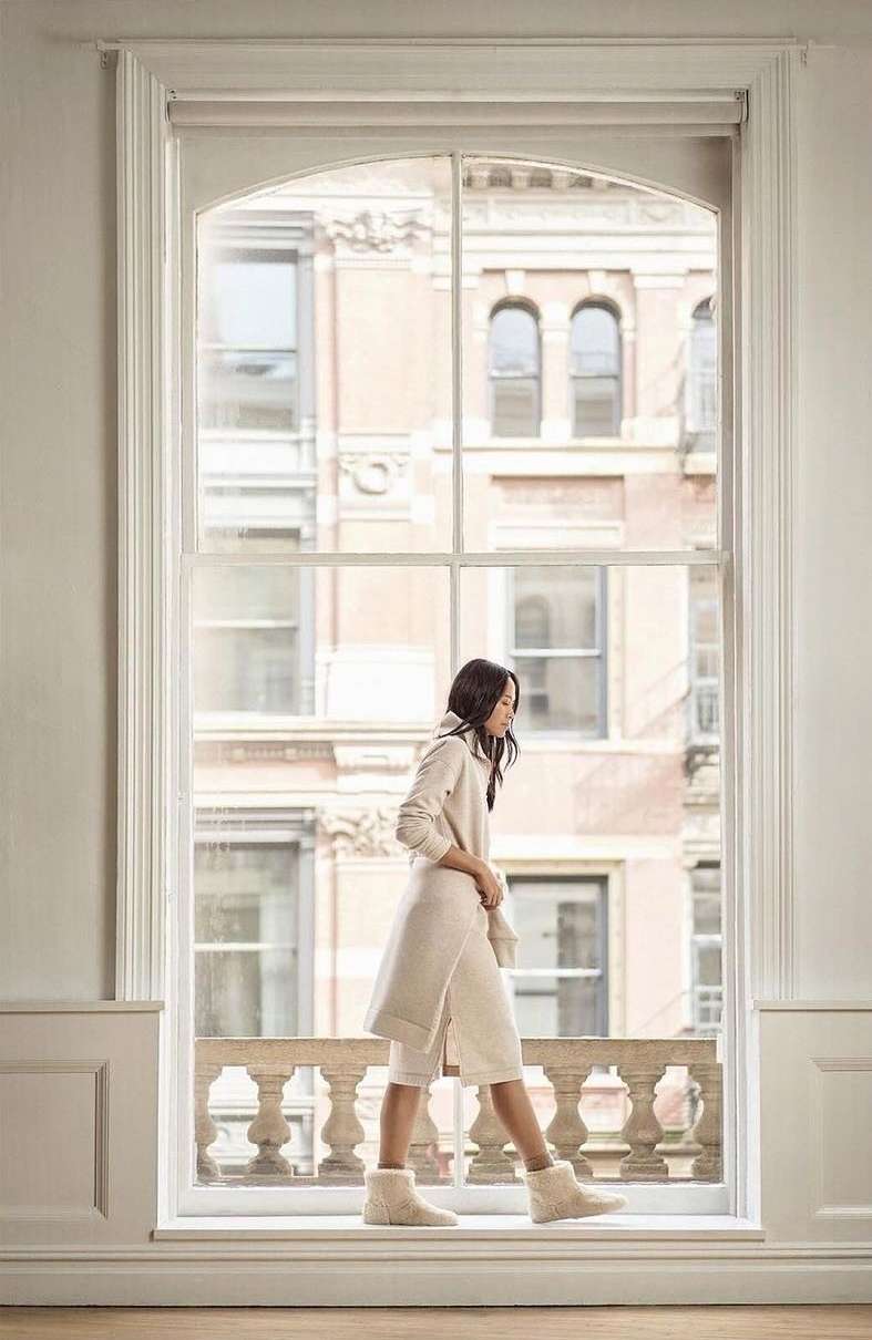 A woman walks along a window ledge in front of a large window with a city view, wearing a hooded sweater and furry boots.