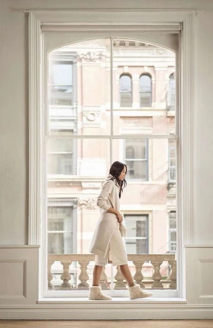 A woman walks along a window ledge in front of a large window with a city view, wearing a hooded sweater and furry boots.