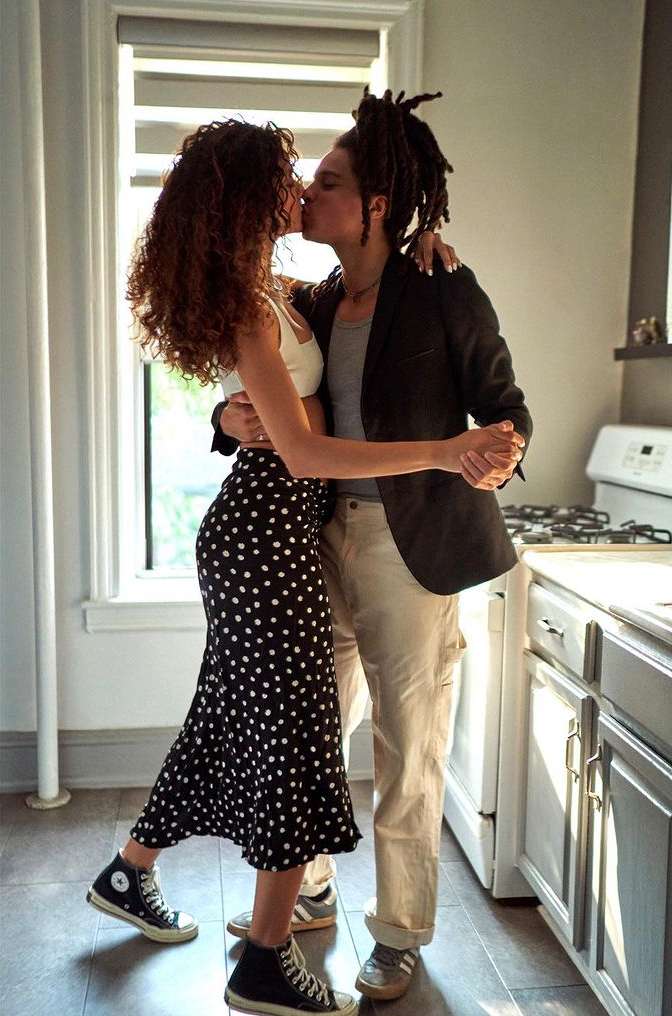 Romantic indoor portrait of a couple kissing while embracing near a bright window, one person wears a blazer and the other a polka dot skirt.