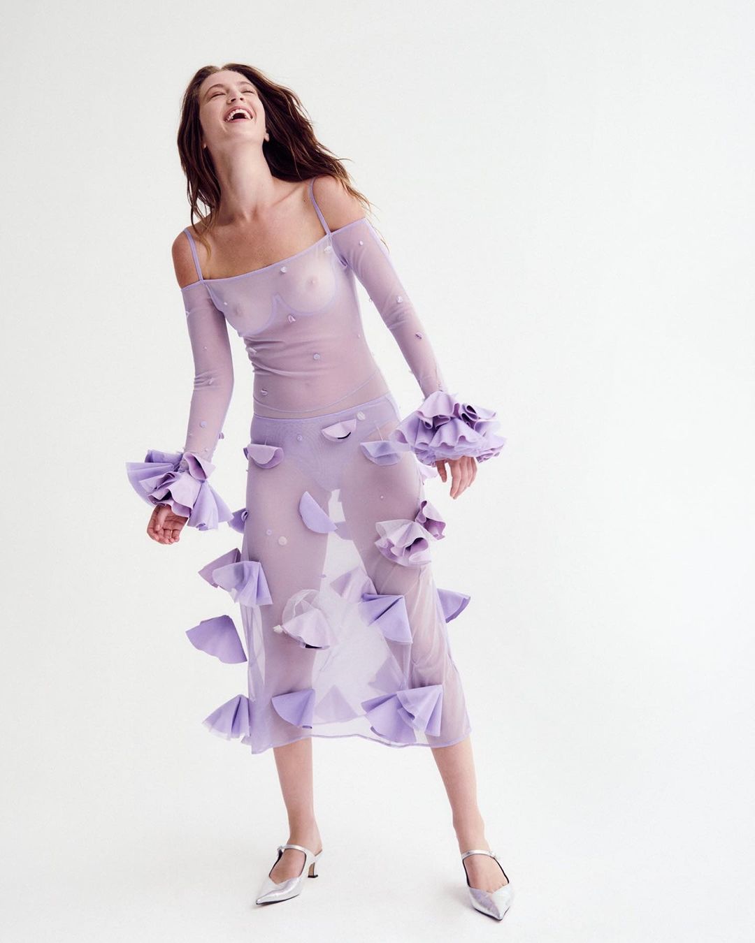 Laughing woman in sheer violet dress with floral accents and silver heels poses in a studio with a bright white background.