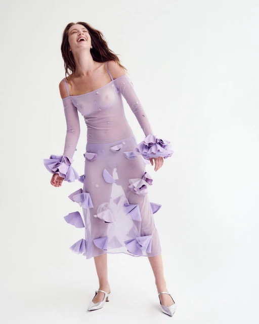 Laughing woman in sheer violet dress with floral accents and silver heels poses in a studio with a bright white background.