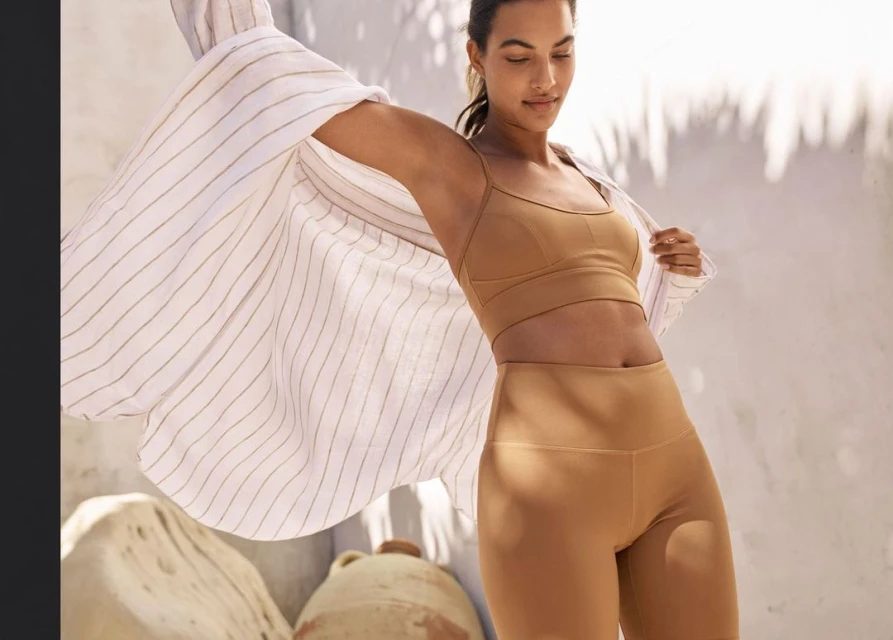 A woman in athletic clothing poses in front of a bright background, wearing a striped shirt, sport bra, and leggings, outdoors.