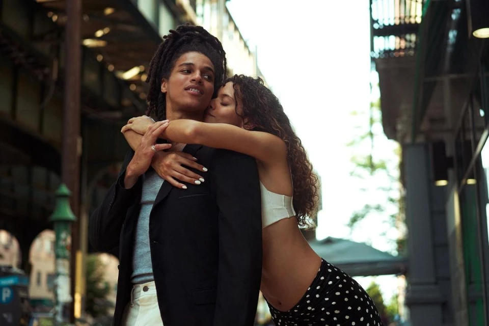 Woman hugs man with dreadlocks on city street. She wears a white top and black polka dot pants, he wears a black blazer.