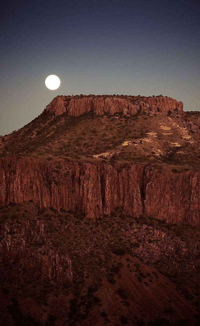 Scenic view of a rocky mountain under a full moon in the night sky, showing rock formations and arid landscape with clear skies.
