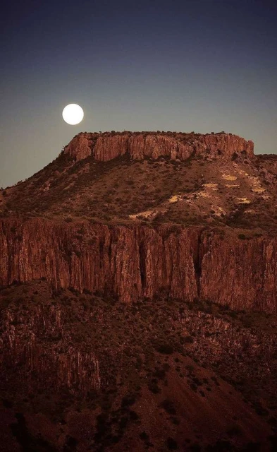 Scenic view of a rocky mountain under a full moon in the night sky, showing rock formations and arid landscape with clear skies.