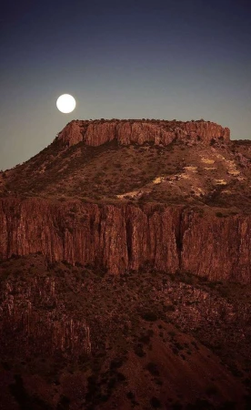 Scenic view of a rocky mountain under a full moon in the night sky, showing rock formations and arid landscape with clear skies.