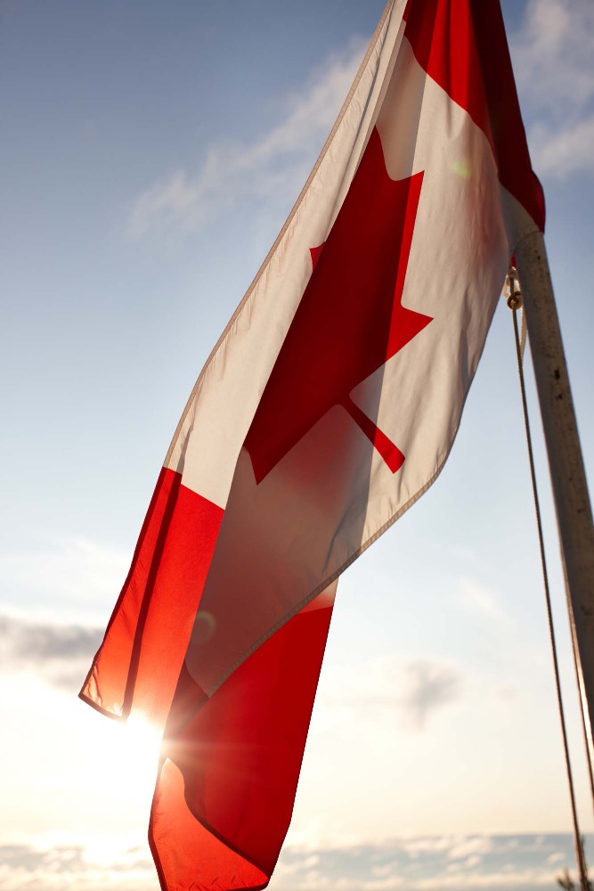 Canadian flag waving against a blue sky with bright sunshine. The red and white flag fills the frame in a patriotic display.