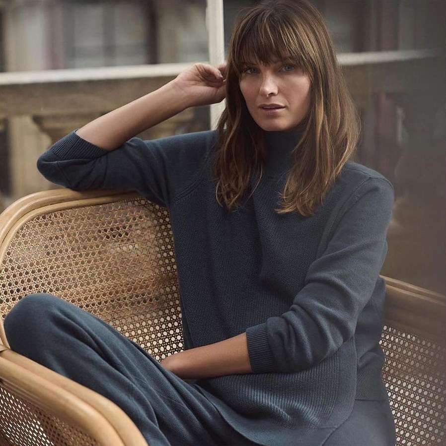 A woman with brown hair sits in a light wicker chair wearing a navy sweater and pants with blurred background.