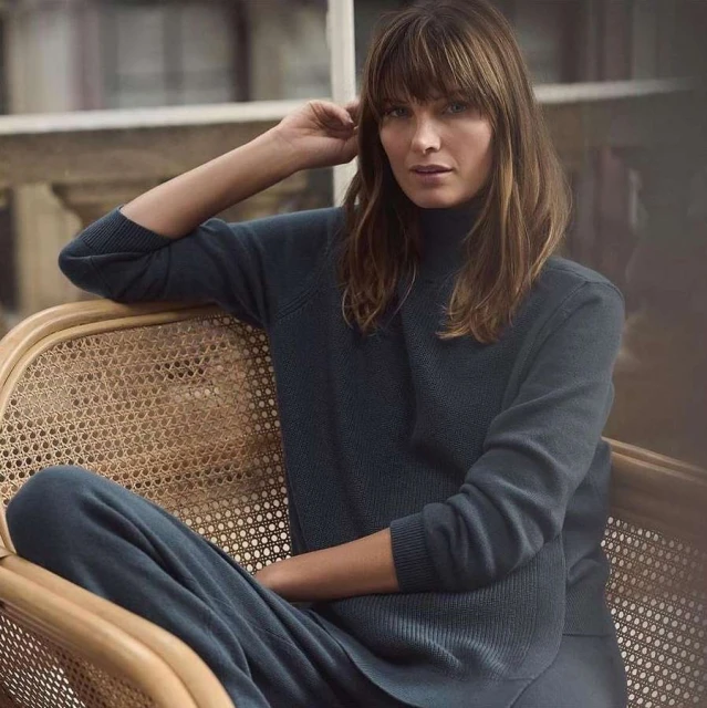 A woman with brown hair sits in a light wicker chair wearing a navy sweater and pants with blurred background.