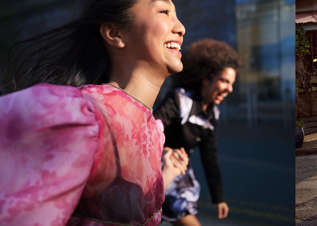 Two women are holding hands and laughing as they walk outdoors in daylight. One woman is wearing a pink patterned top.