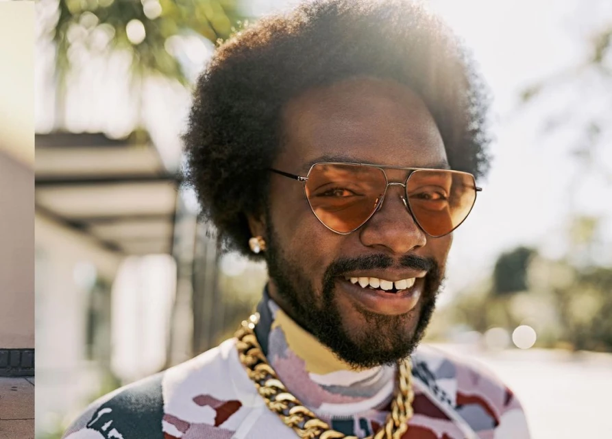 Close-up of an adult male with sunglasses and a chain, smiling outdoors with sun flare and foliage in the soft-focus background.