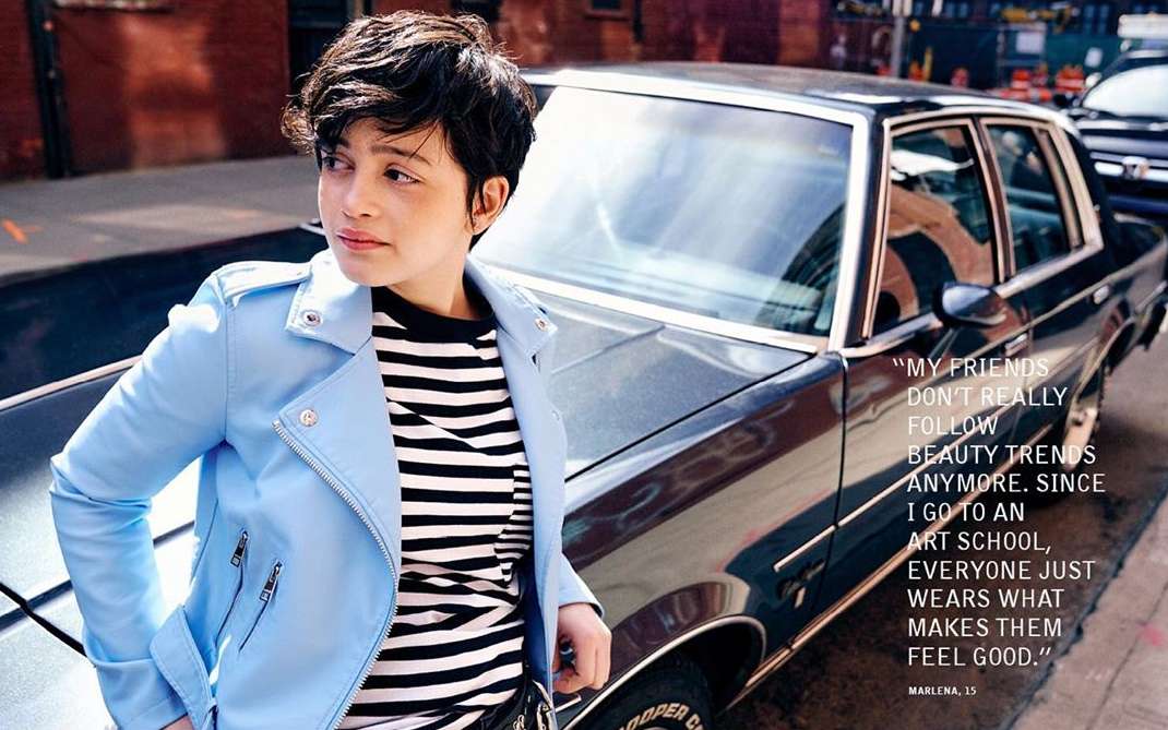 Stylish adult with short dark hair stands by vintage car on city street wearing striped top and blue moto jacket in bright sunlight.
