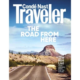 An empty desert highway curving toward a distant rocky mountain under a bright cloudy sky, featured on a travel magazine cover.