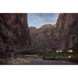 Campsite by a river in a steep canyon with glowing tents and a campfire under an overcast sky.
