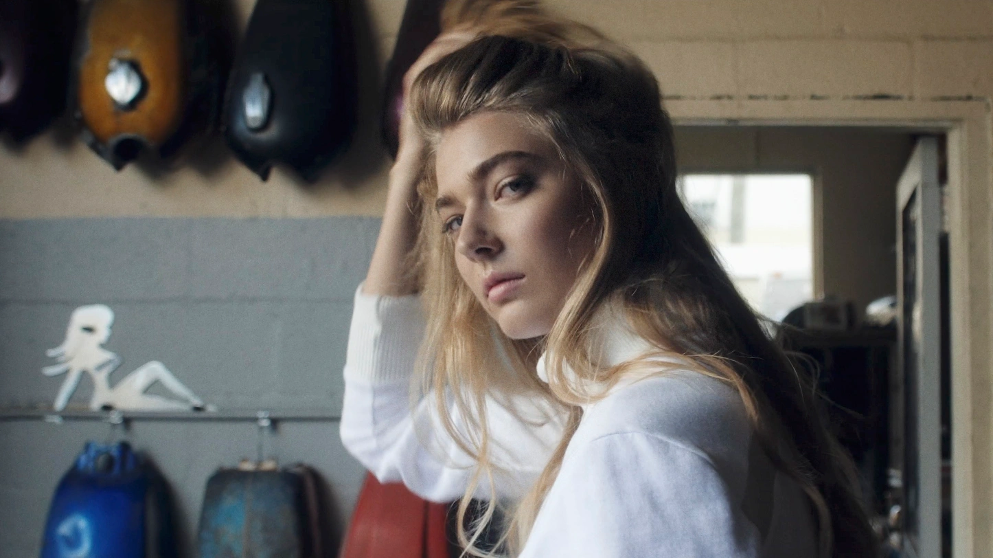 A woman with long blonde hair wearing a white turtleneck sweater posing in a workshop with motorcycle parts hanging on the wall.