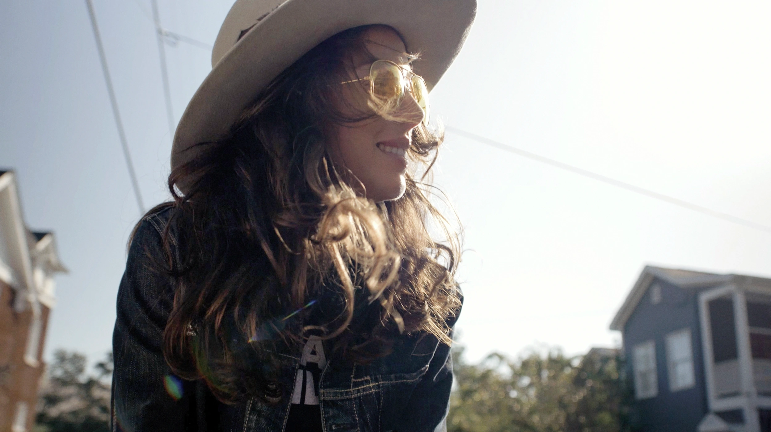 Woman wearing yellow tinted sunglasses and a wide brimmed hat outdoors by houses
