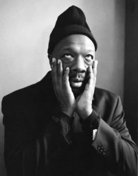 Black and white portrait of a man wearing a beanie and dark suit, holding his face in his hands with eyes rolling upward.