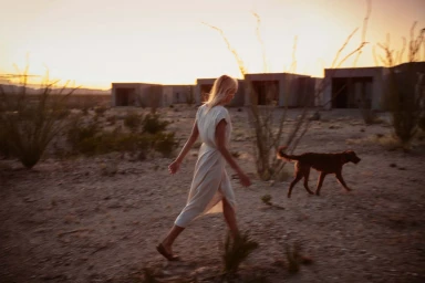 A blonde woman in a cream dress walks through a desert landscape with a dog at sunset, photographed by Squire Fox.