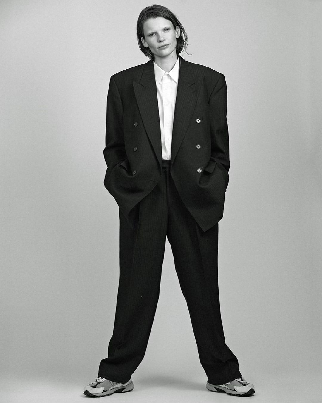 Woman in oversized black suit and white shirt stands in a photo studio with a neutral grey background, wearing grey running shoes.