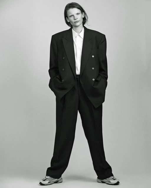 Woman in oversized black suit and white shirt stands in a photo studio with a neutral grey background, wearing grey running shoes.