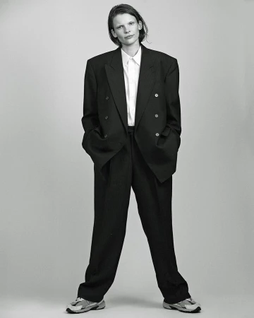 Full length portrait of a model in an oversized black suit and white shirt against a neutral grey background