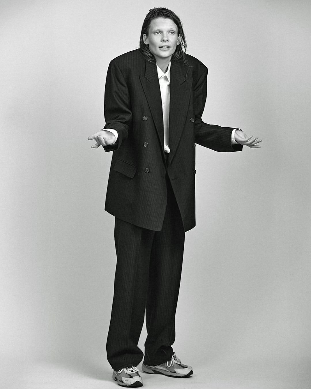 Monochrome full shot of an adult posing in an oversized pinstripe suit with a white shirt and sneakers against a plain background.