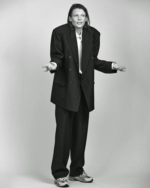 Monochrome full shot of an adult posing in an oversized pinstripe suit with a white shirt and sneakers against a plain background.