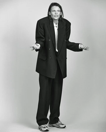 Monochrome full shot of an adult posing in an oversized pinstripe suit with a white shirt and sneakers against a plain background.