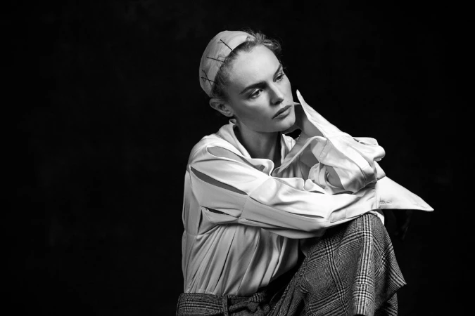 Monochrome portrait of a woman wearing a white collared shirt and patterned trousers against a dark background.