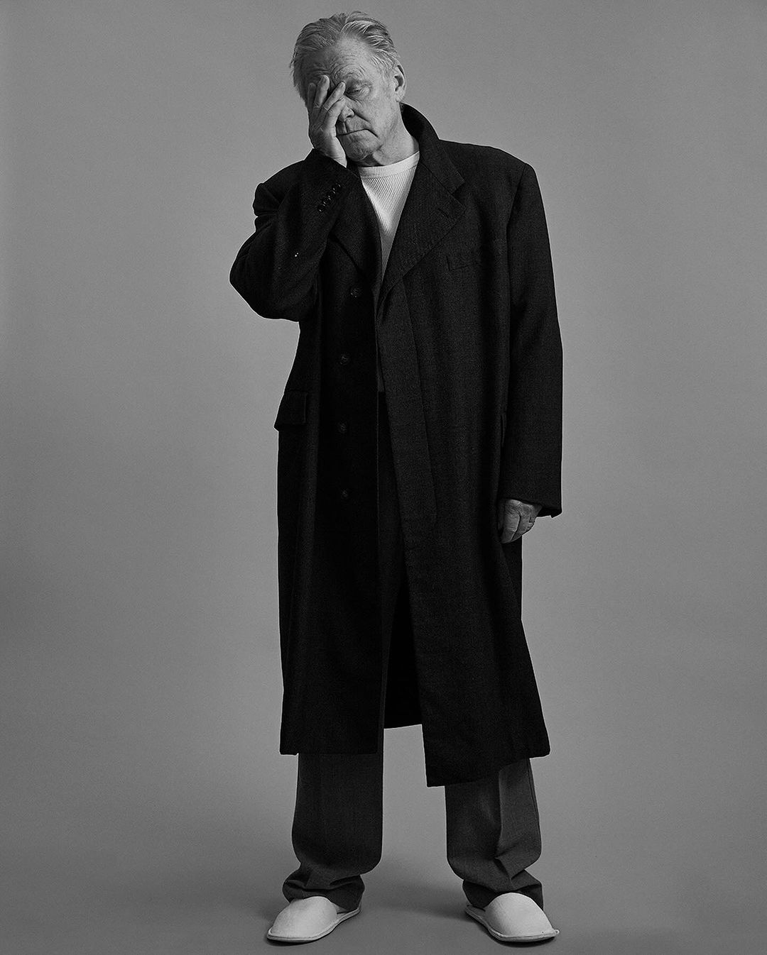 An elderly male in a black overcoat stands indoors with his hand covering his face, set against a simple grey backdrop in soft lighting.