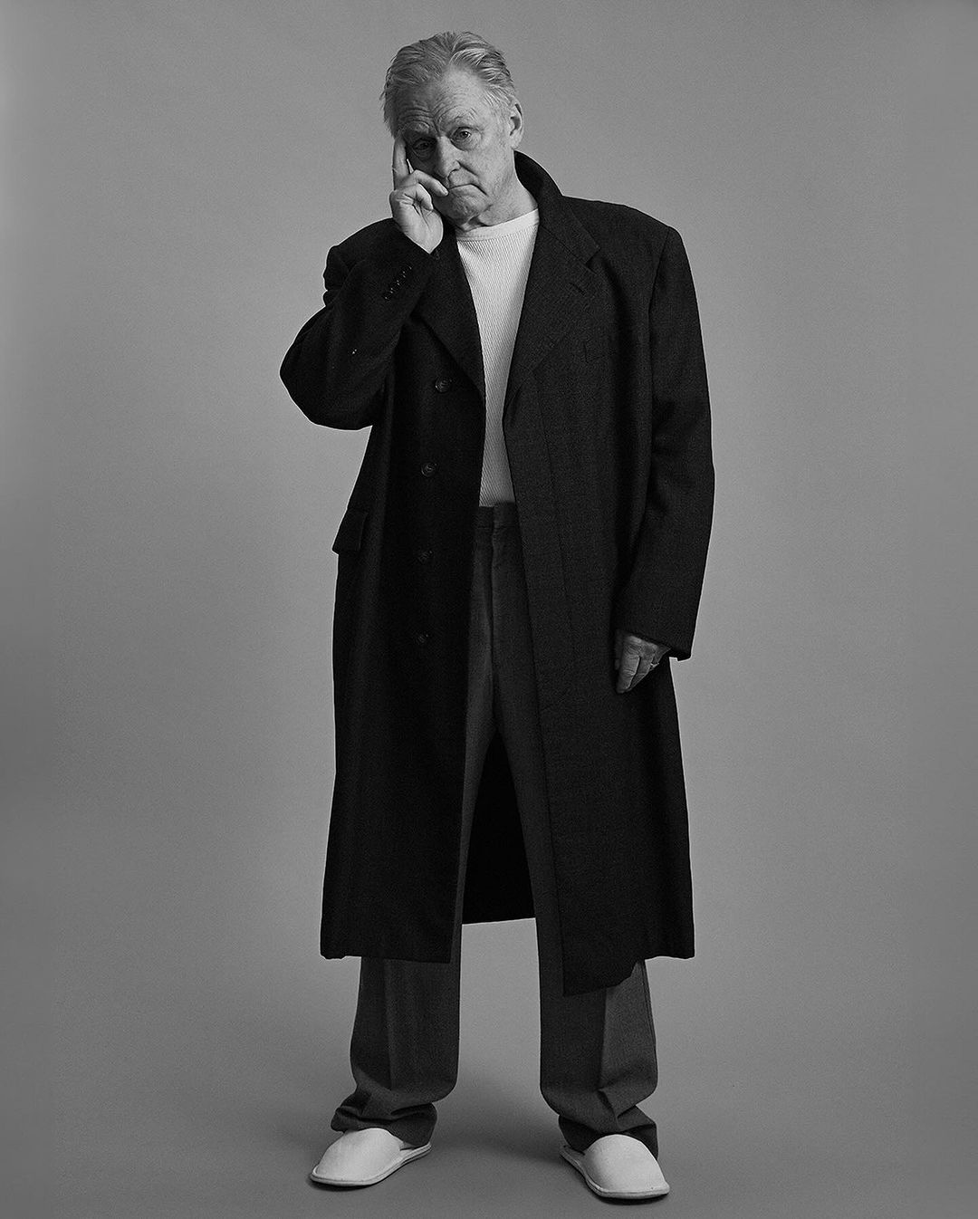 Black and white portrait of an adult male in a long coat, white shirt, trousers, and slippers standing indoors with hand on head.