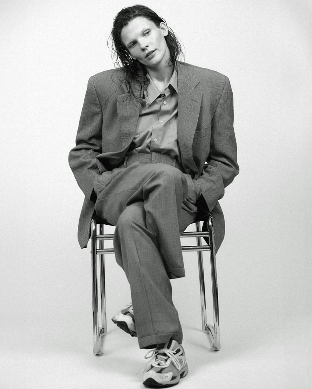 Model with wet hair in an oversized grey suit sitting on a chair against a white seamless background